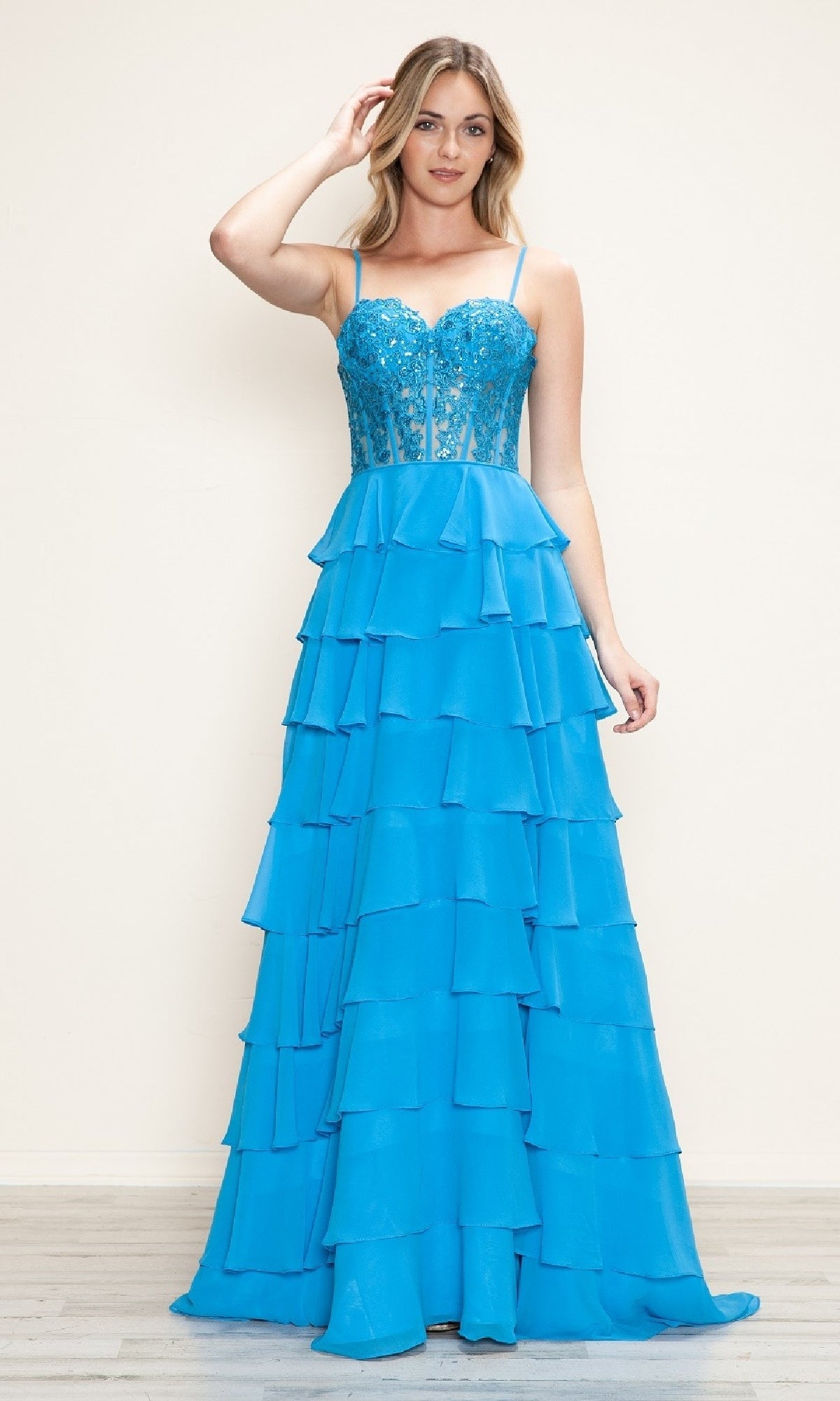 A woman wears the Long Tiered Corset Prom Dress: Poly USA 9698, featuring a bright blue floor-length design with a sequined corset-style bodice, thin straps, and a tiered ruffled skirt.